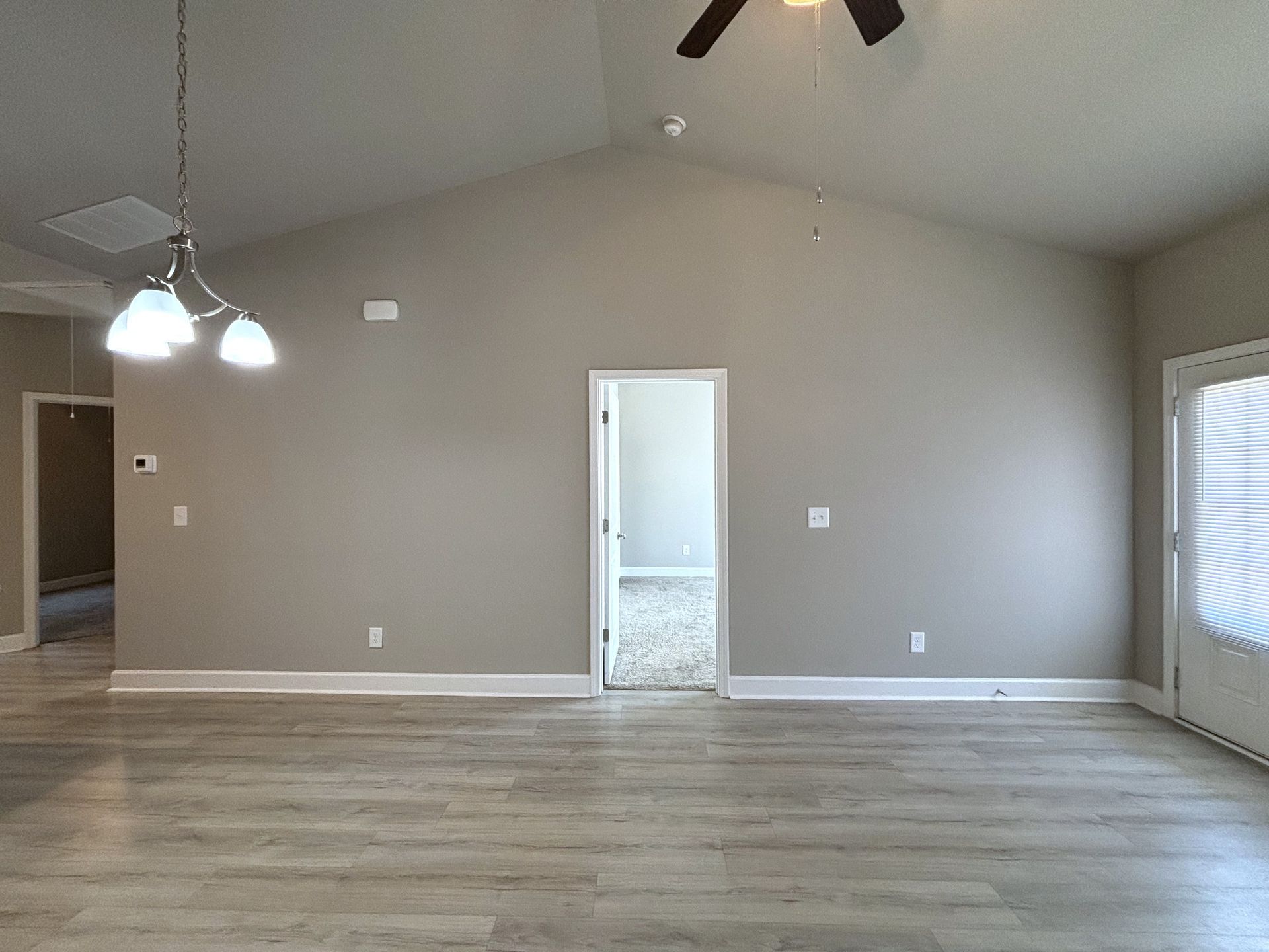 Empty beige living room with wood floors, ceiling fan, chandelier, and a doorway to a tiled room