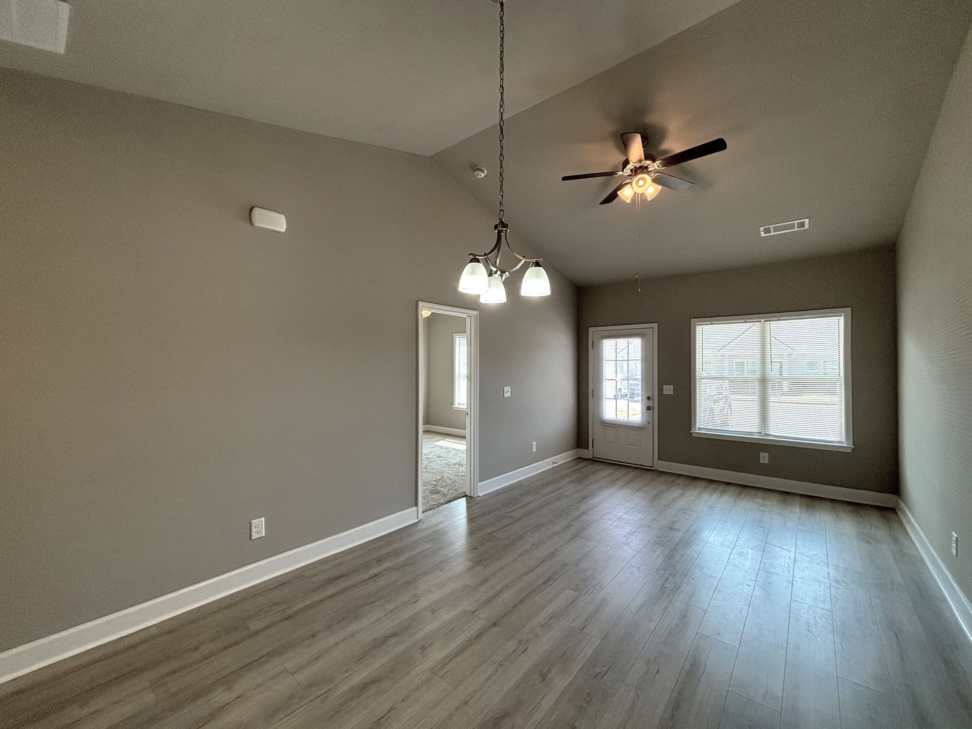 Empty beige living room with gray wood floors, ceiling fan, and bright windows and door letting in sunlight