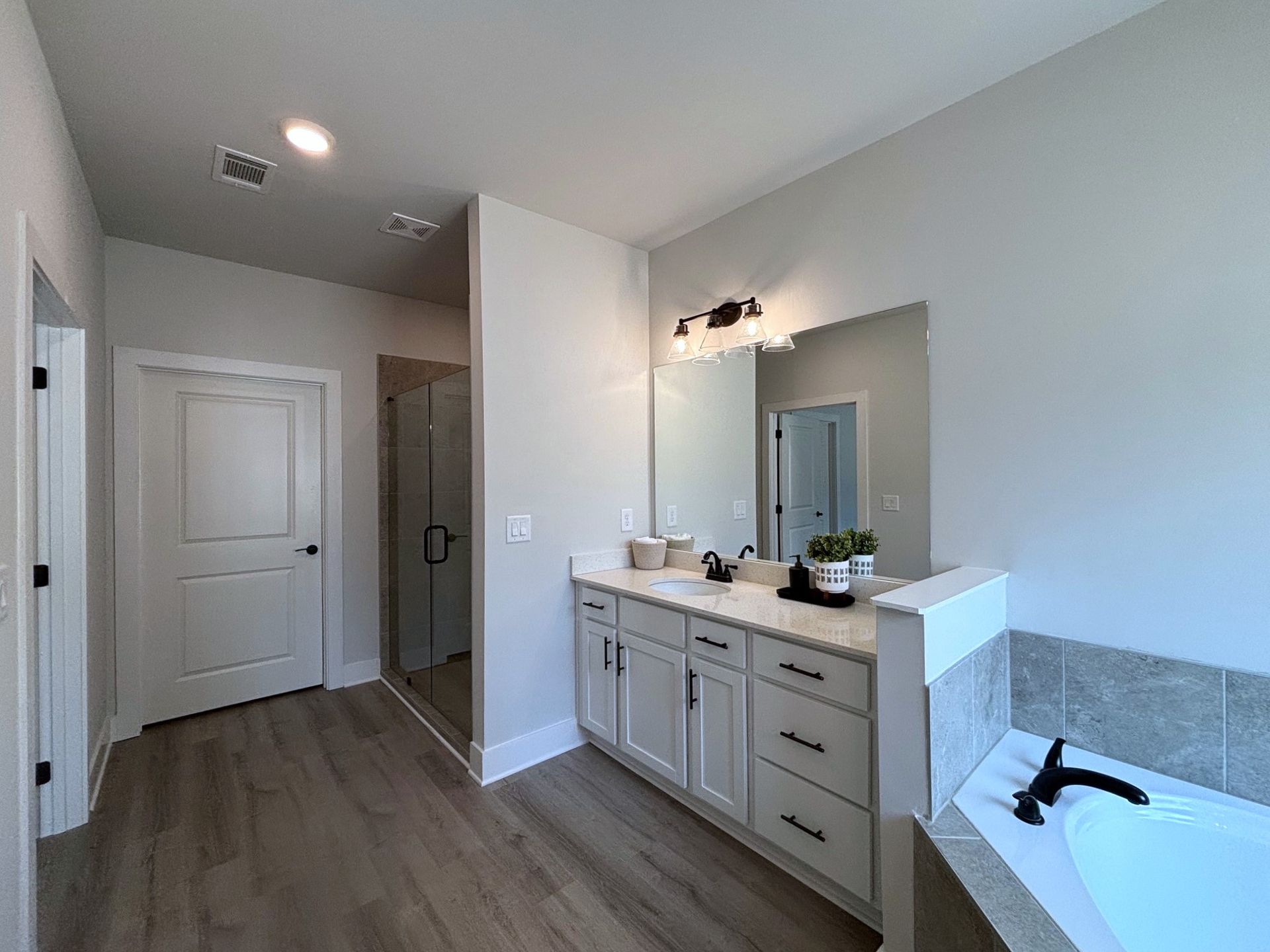 Bathroom with double vanity, white walls, gray cabinets, mirror lights, and a tub in the corner.