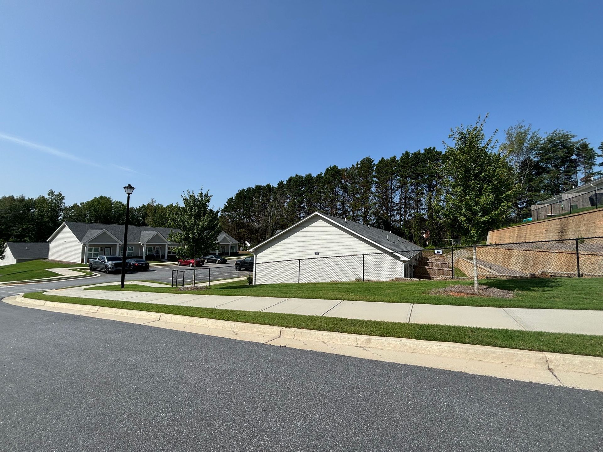 Quiet suburban street with gray houses, sidewalk, green lawns, and trees under a clear blue sky