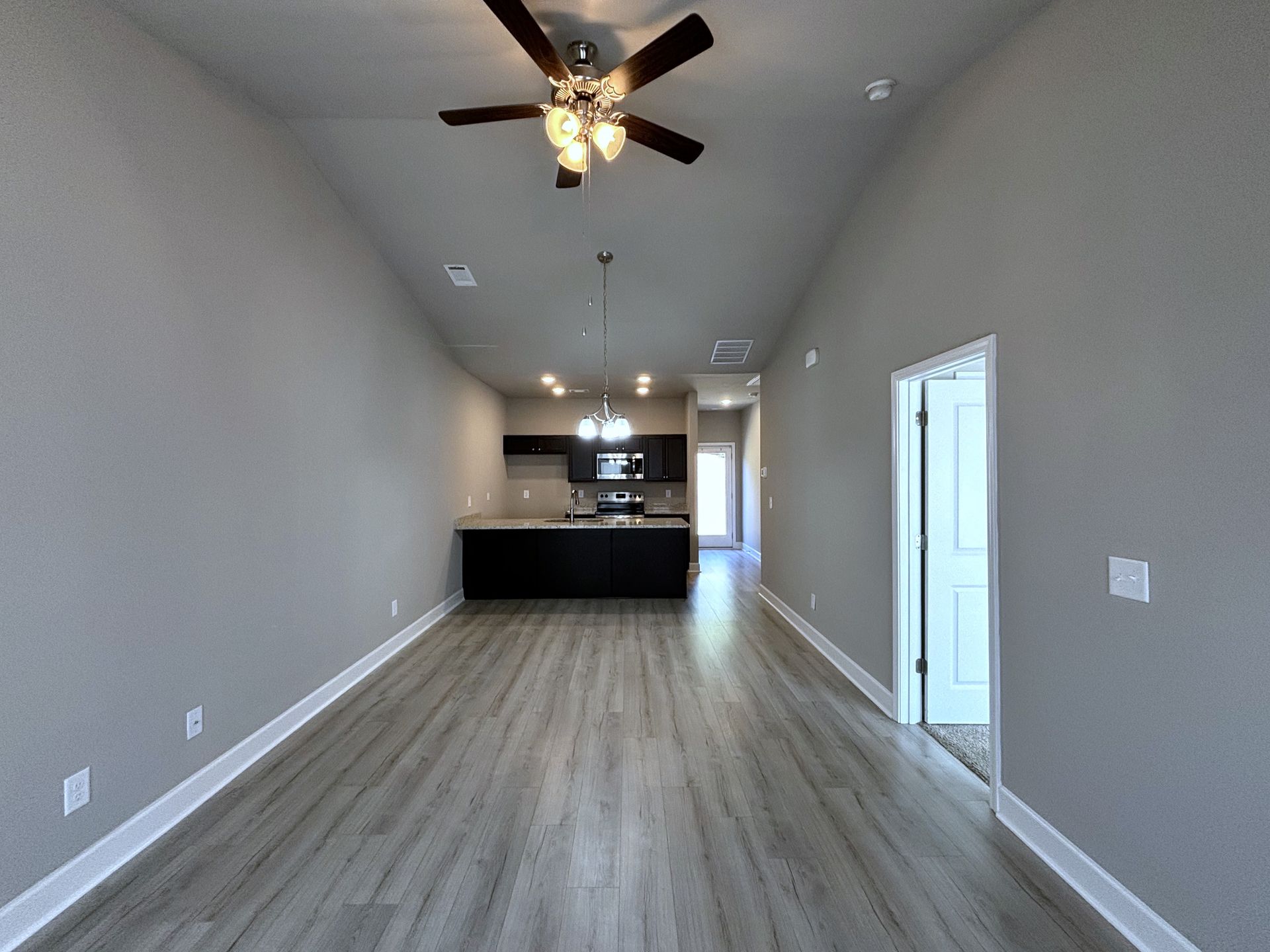 Empty beige living room with vaulted ceiling, ceiling fan, and open kitchen at the back.