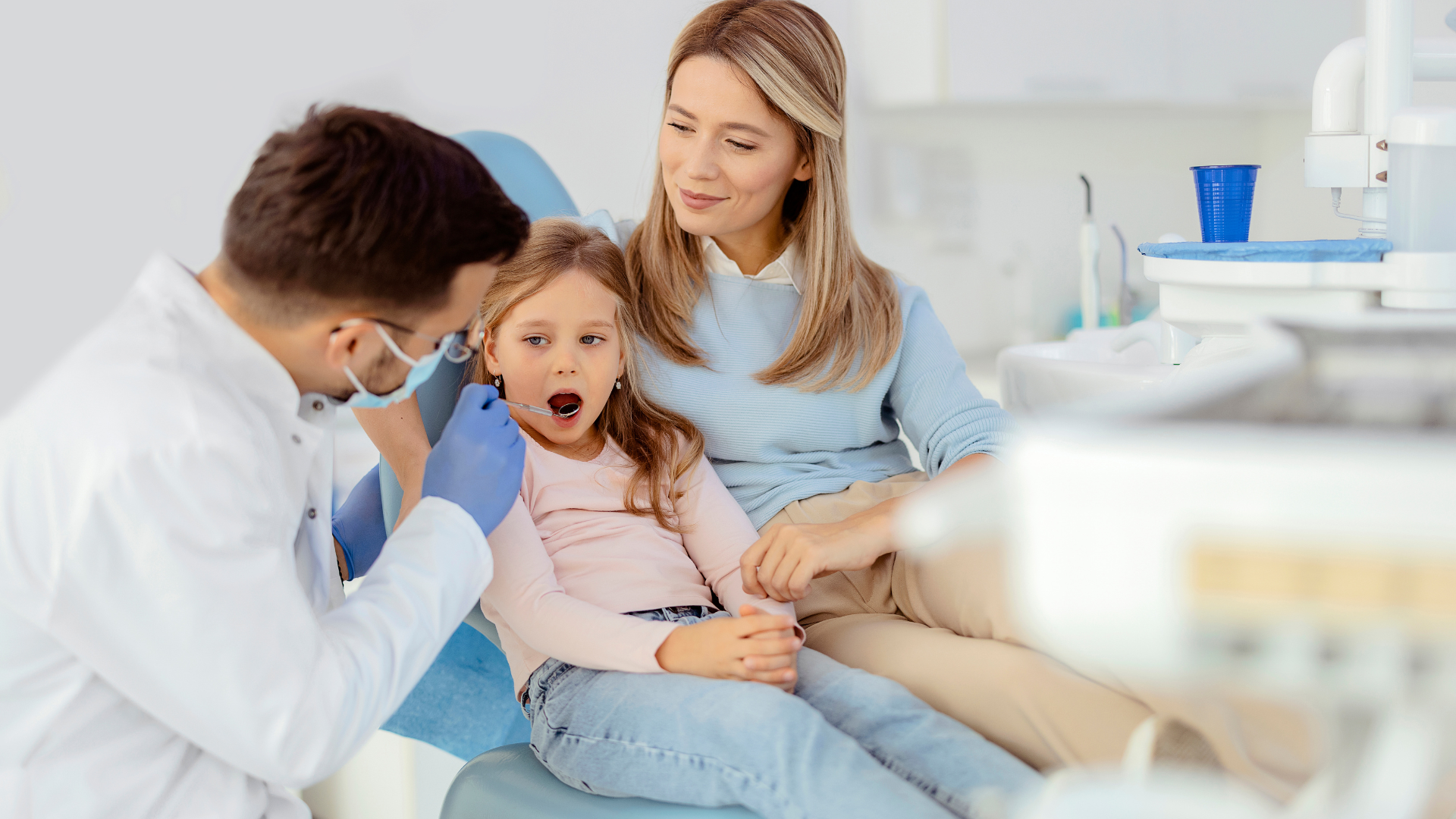 A little girl is sitting in a dental chair with her mother while a dentist examines her teeth.