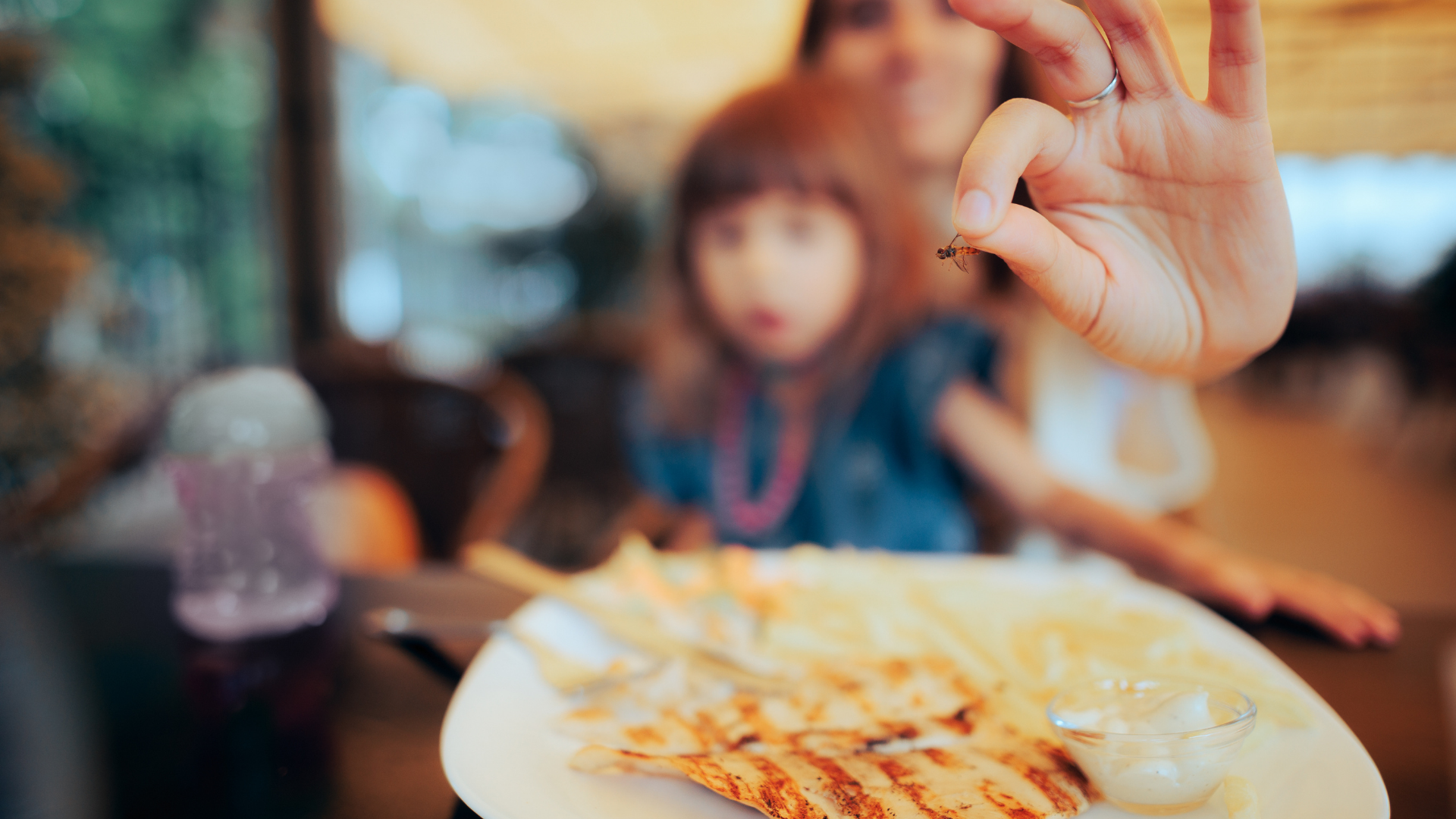 A woman is holding a small bug in her hand in front of a plate of food.