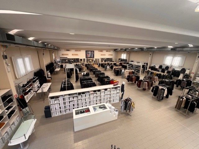 Ampio interno di un negozio di abbigliamento con file di scaffali, scatole ed espositori. Pareti beige, luci intense.