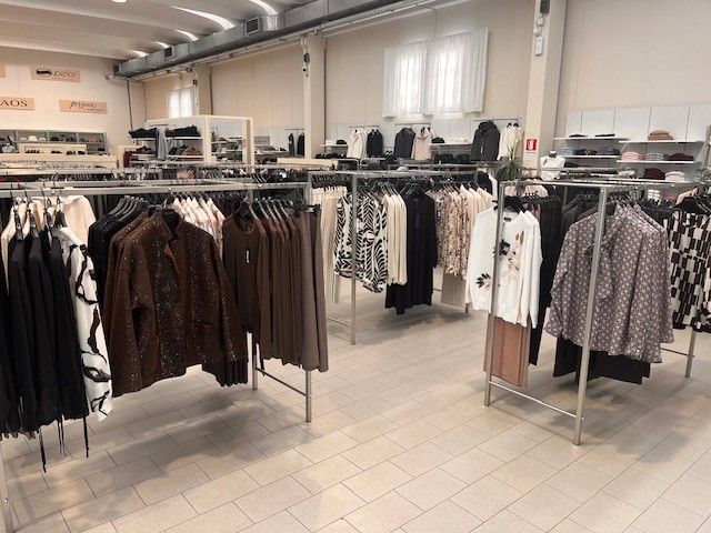 Interno di un negozio di abbigliamento con scaffali pieni di vestiti, palette di colori neutri.
