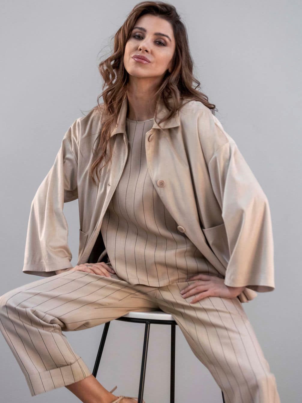 Donna in abito color cuoio, seduta su uno sgabello, con le mani sui fianchi, che guarda l'osservatore.