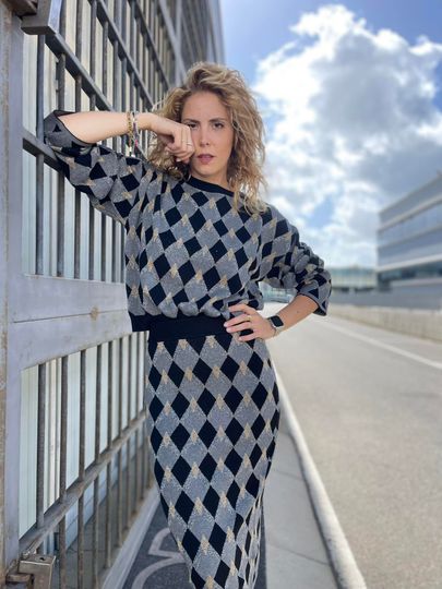 Donna con maglione a rombi e gonna, in posa vicino a una recinzione, all'aperto.