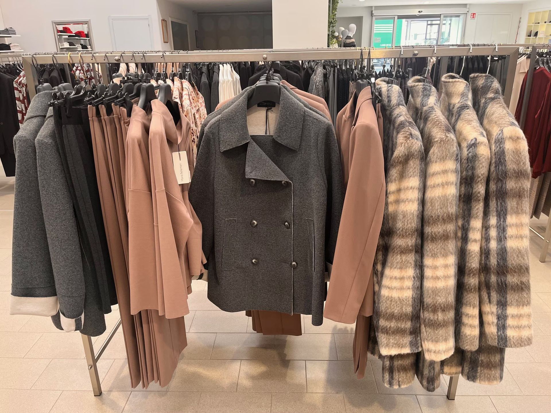 L'espositore per abiti di un negozio espone cappotti e pantaloni nelle tonalità del grigio, del marrone chiaro e del marrone.