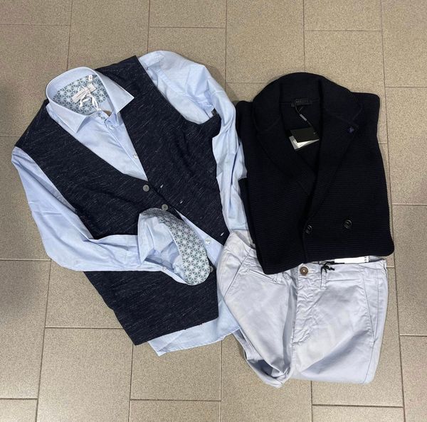 Abbigliamento maschile: camicia blu, gilet blu navy, giacca nera e pantaloni azzurri su un pavimento piastrellato.