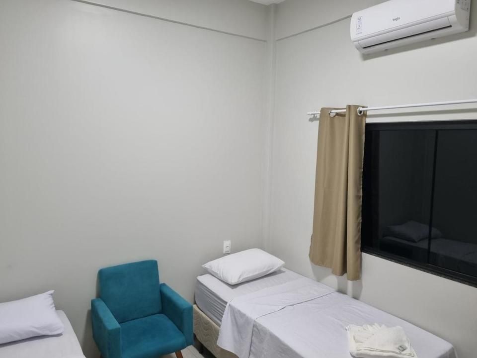 Um pequeno quarto com duas camas de solteiro, uma poltrona azul e uma janela com cortina bege.