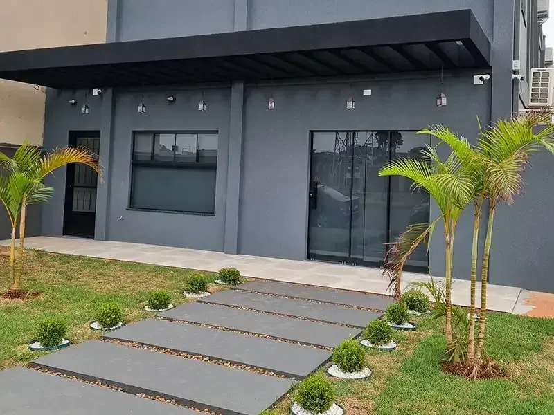 Prédio cinza com toldo e caminho pretos; palmeiras e arbustos na frente.