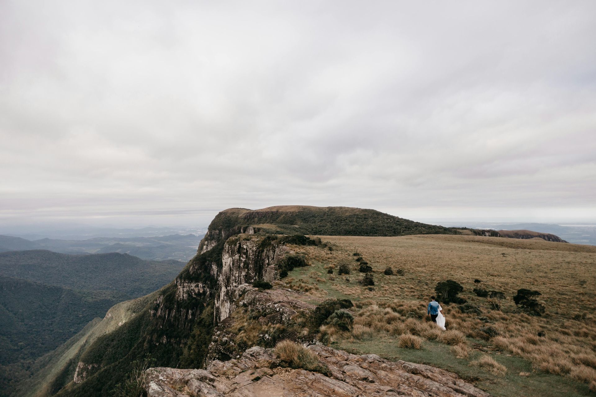 Casal na beira de um penhasco com uma vasta paisagem sob um céu nublado.
