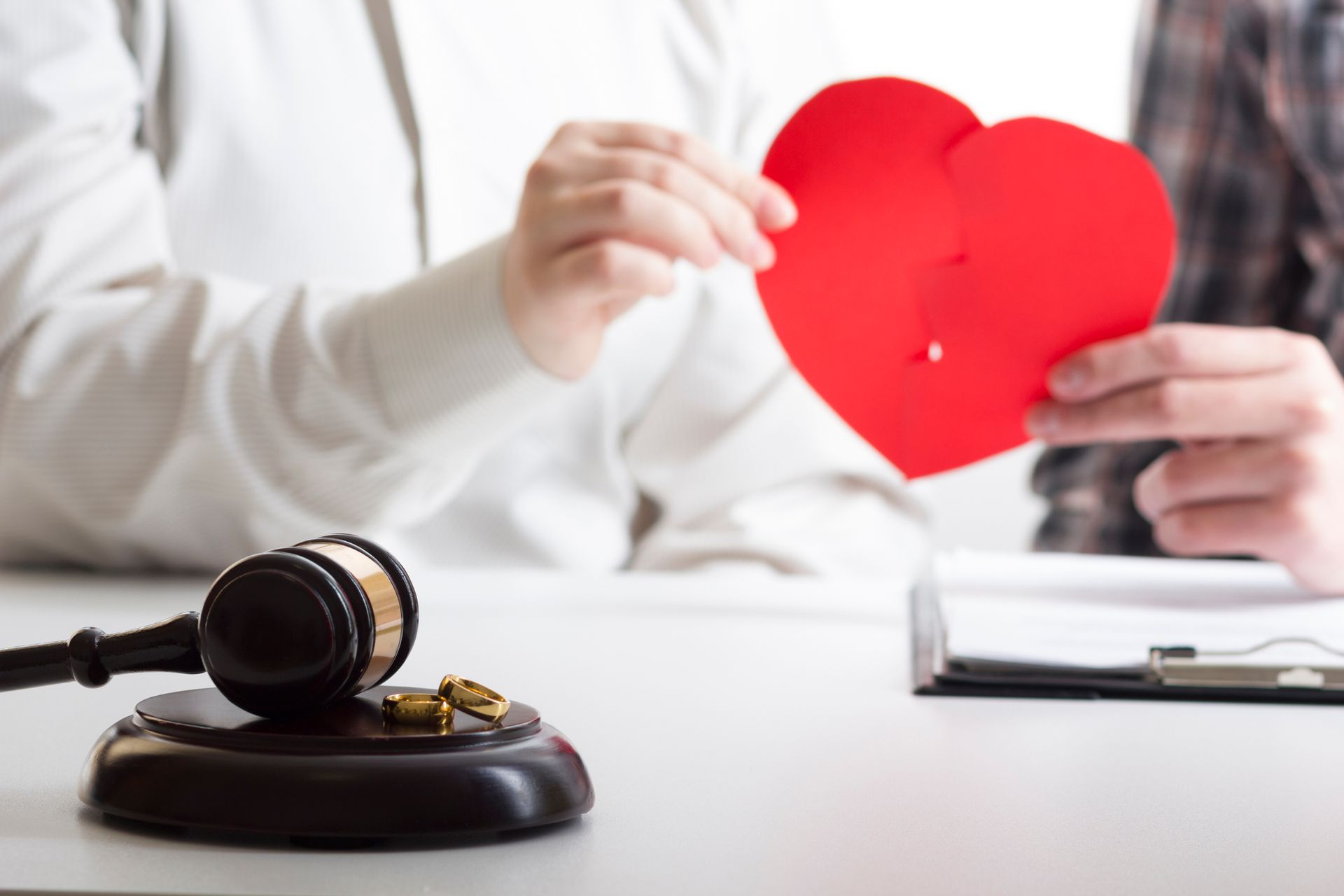Hands of two people hold a cut paper heart, behind a wooden base with a gavel and two wedding rings.