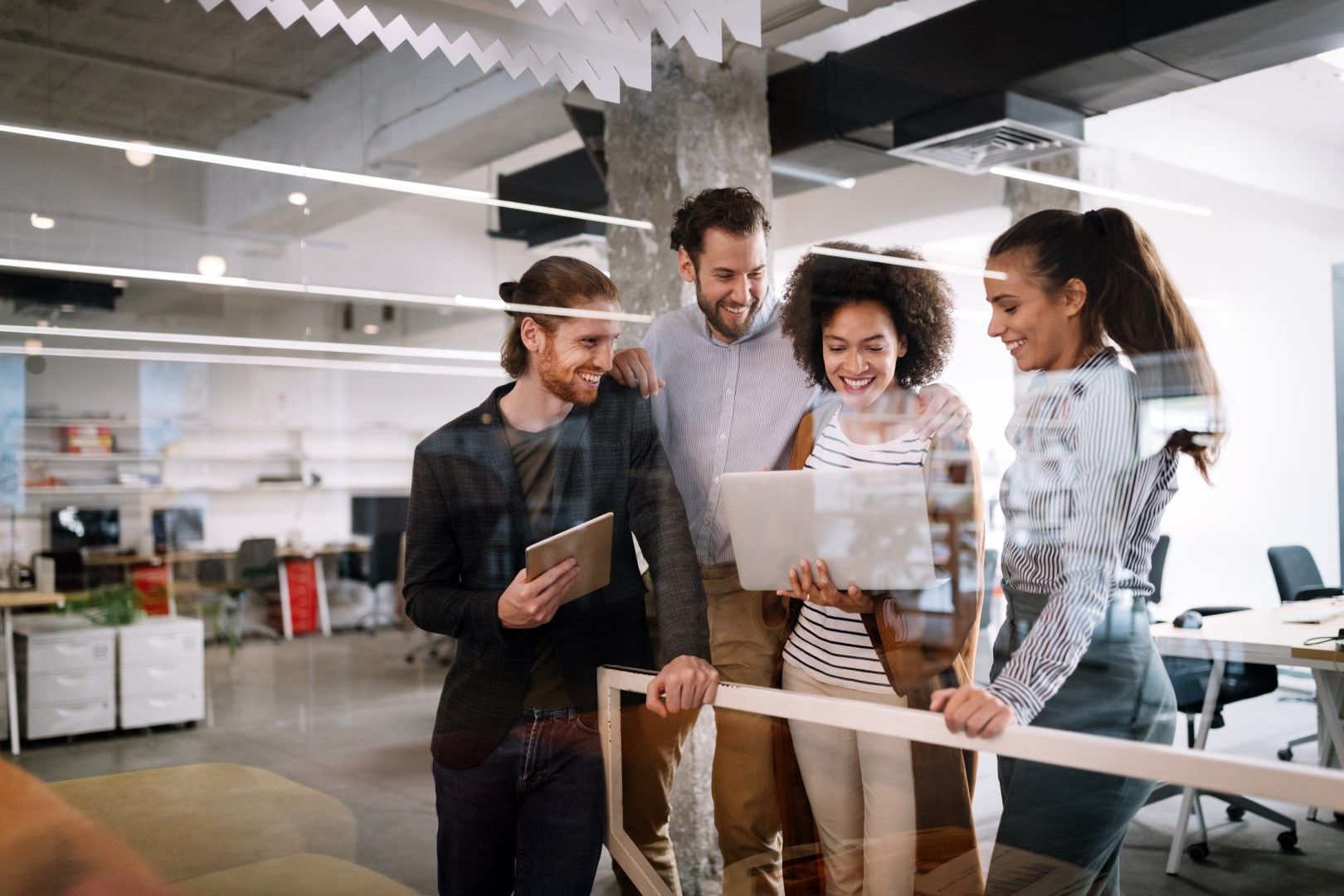 Group of diverse office workers looking at a laptop, smiling, in a modern office setting.