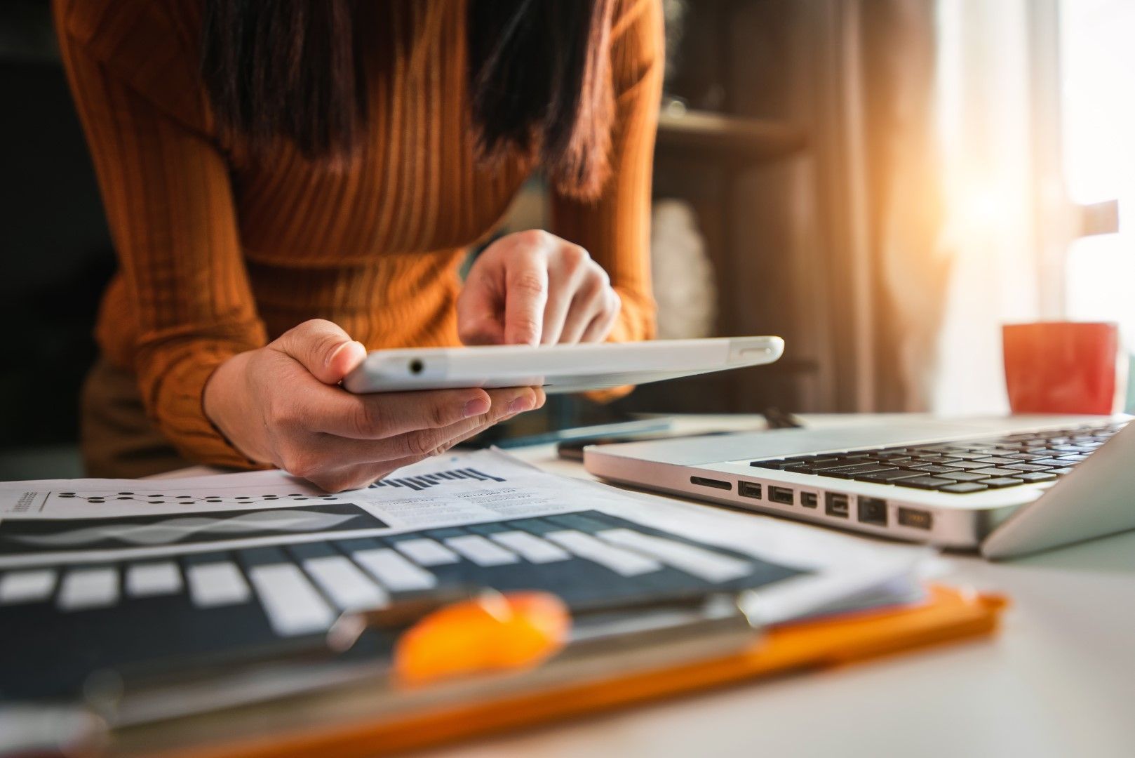 Woman working on tablet at desk with laptop and financial charts.