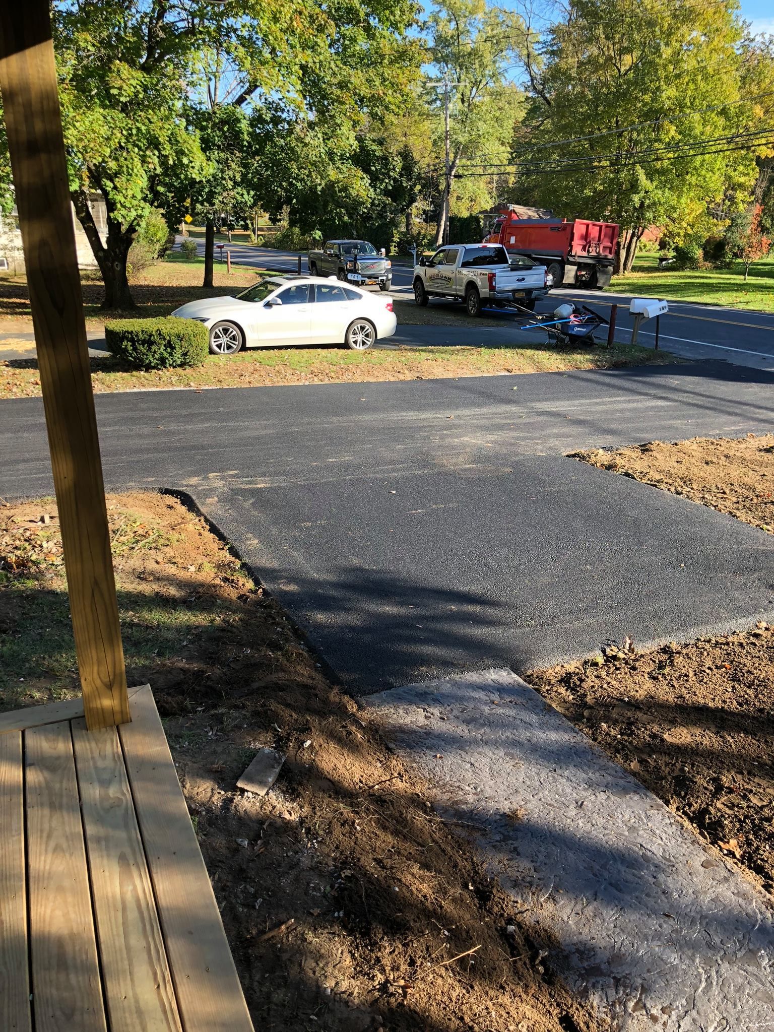 New asphalt driveway under construction; dirt edges; white car and trucks in the background.