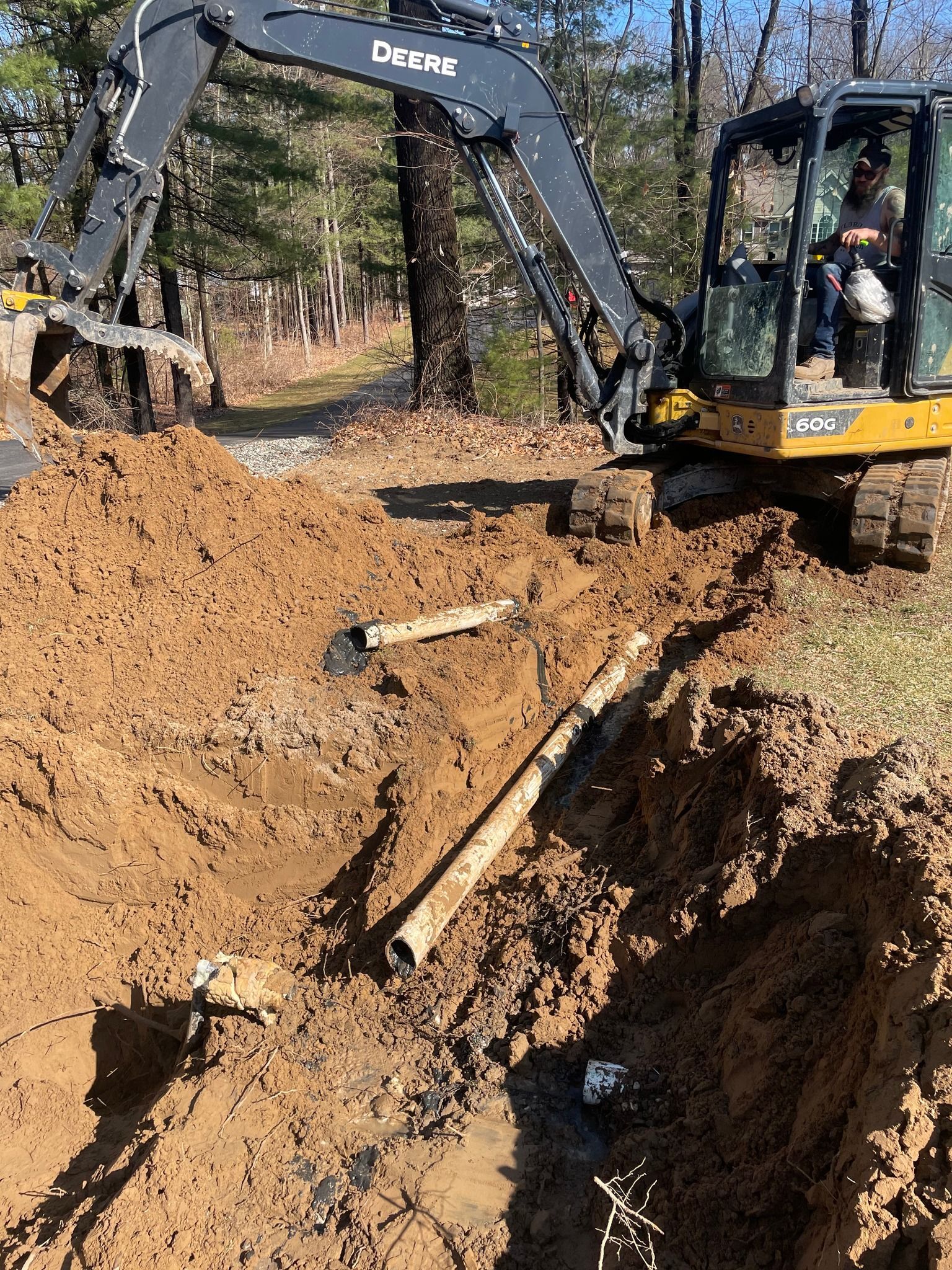 Excavator digging in muddy ground; exposed pipes visible. Operator in cab.