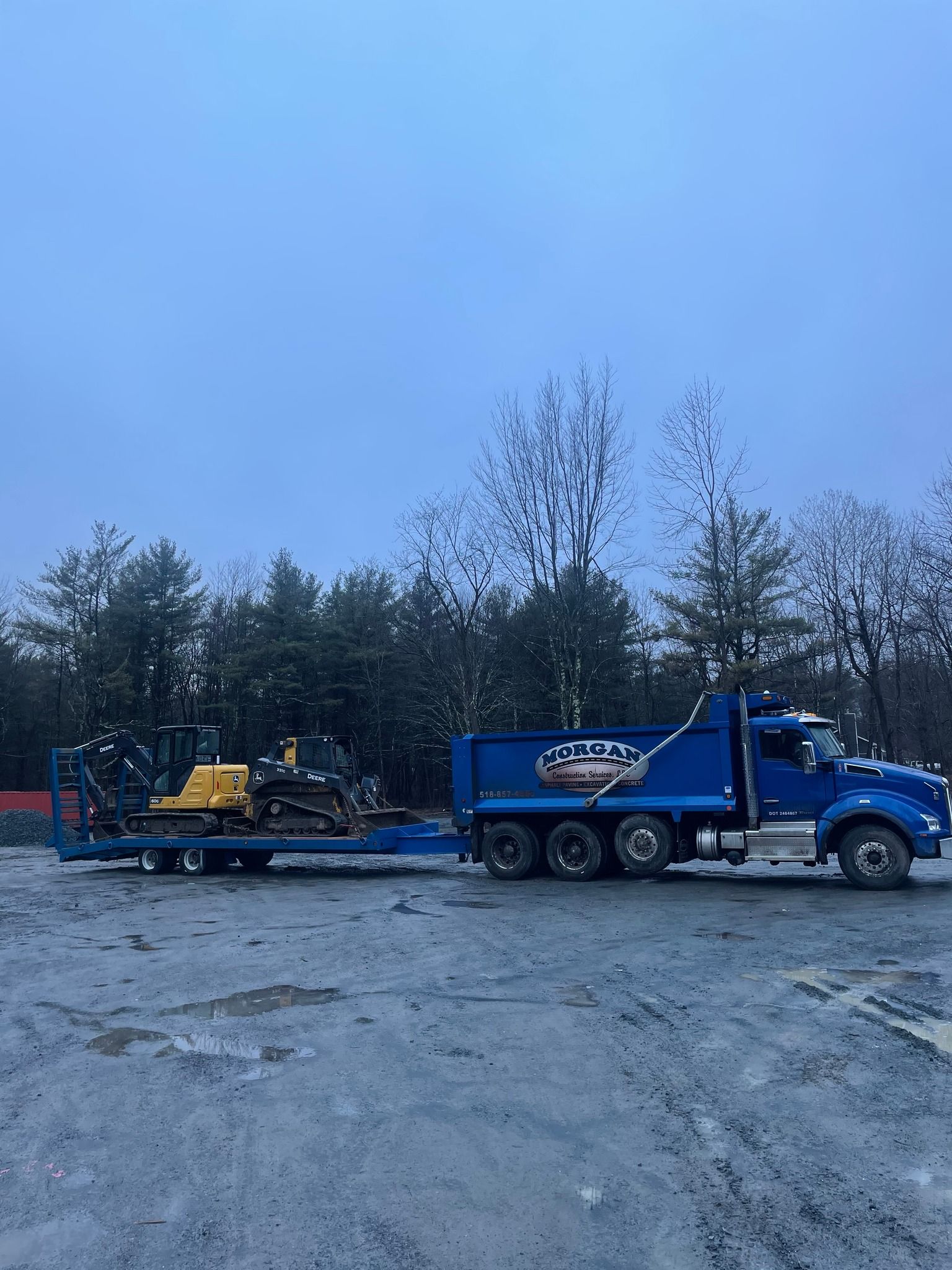 Blue truck towing a trailer with a yellow construction vehicle, parked on gravel, with trees in the background under a cloudy sky.