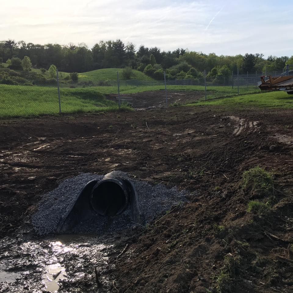 A large black pipe in a muddy construction site. Green hills and trees are in the background.