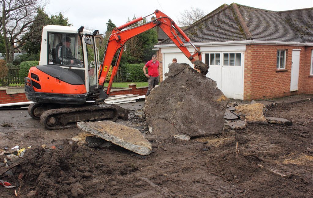 Orange excavator, person operating it, digging up debris near a brick building.