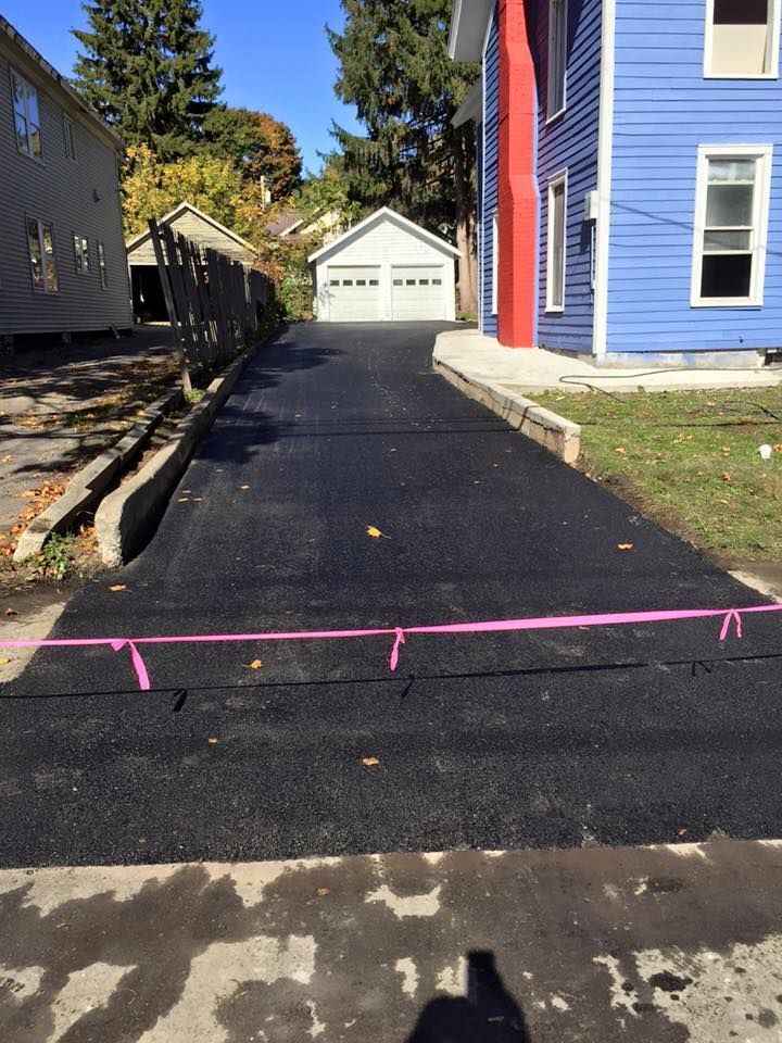 Asphalt driveway leading to a garage. Pink tape lines edges. Blue house on right, other houses in the background.