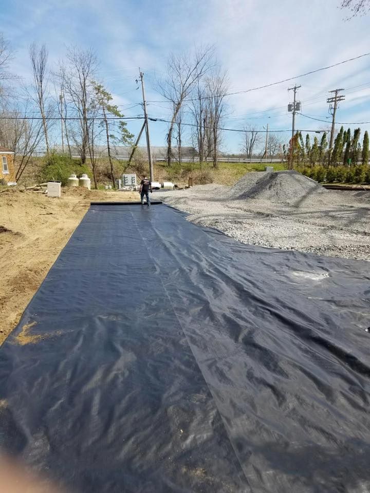 Person installing black landscape fabric, next to a gravel pile, in an outdoor setting.