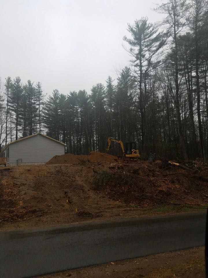 Excavator working on a dirt mound next to a white building; trees in background. Overcast day.
