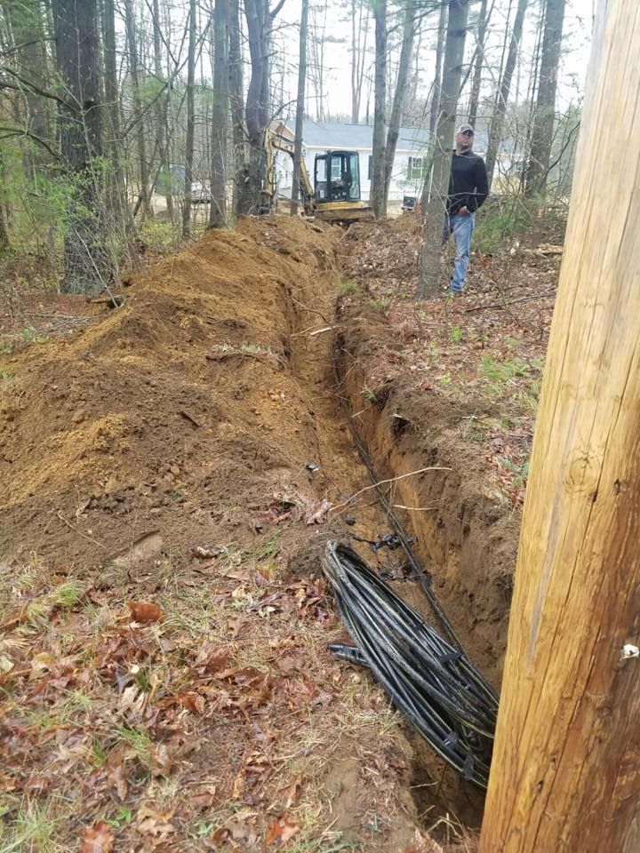 Trench dug through a wooded area for buried cable. A man and small excavator are visible.