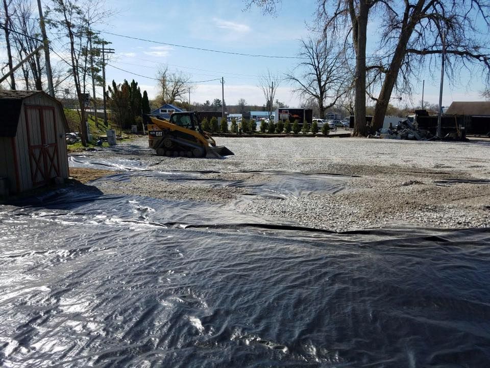 Construction site with a skid steer loader on a gravel lot covered by black plastic; trees in the background.