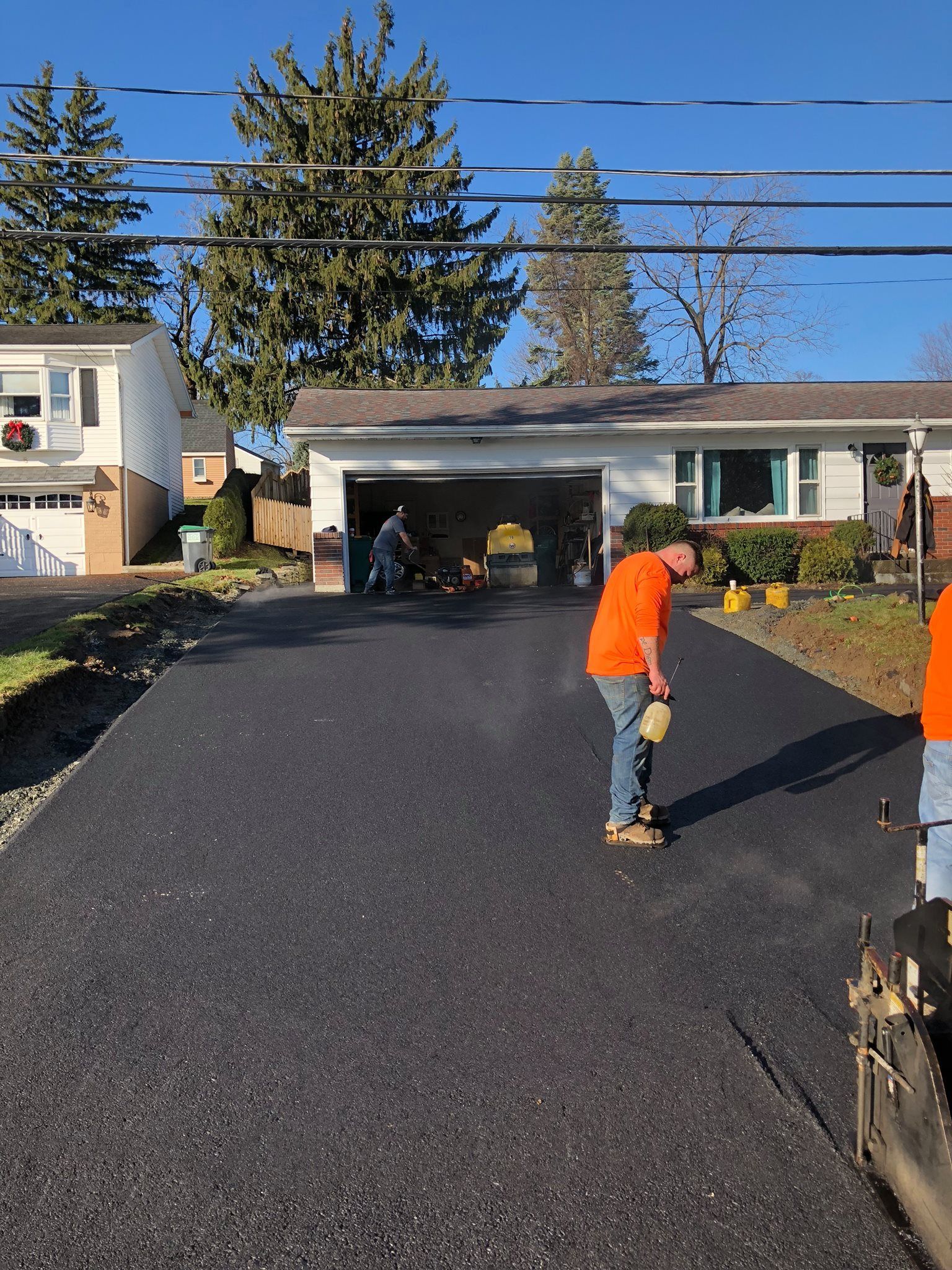 Workers paving a residential driveway; asphalt, garage, trees, blue sky.