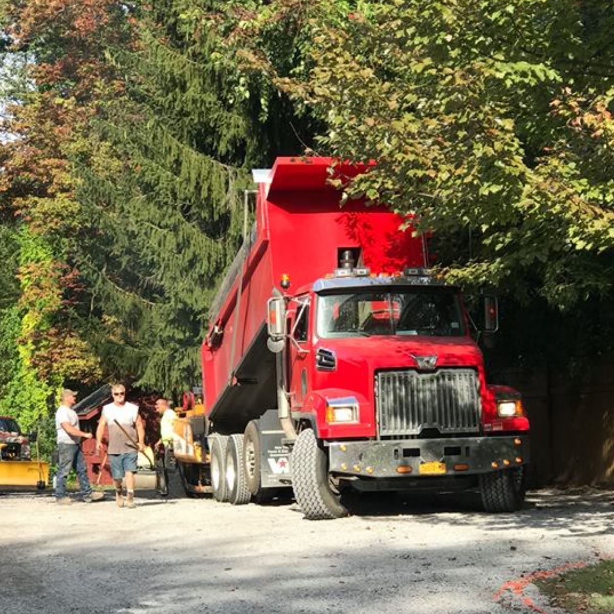 Red dump truck unloading on a road near a group of workers and trees.