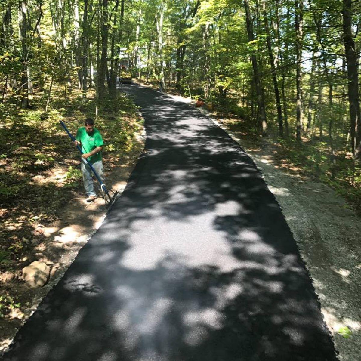 Man in green shirt sprays black asphalt path winding through a forest.