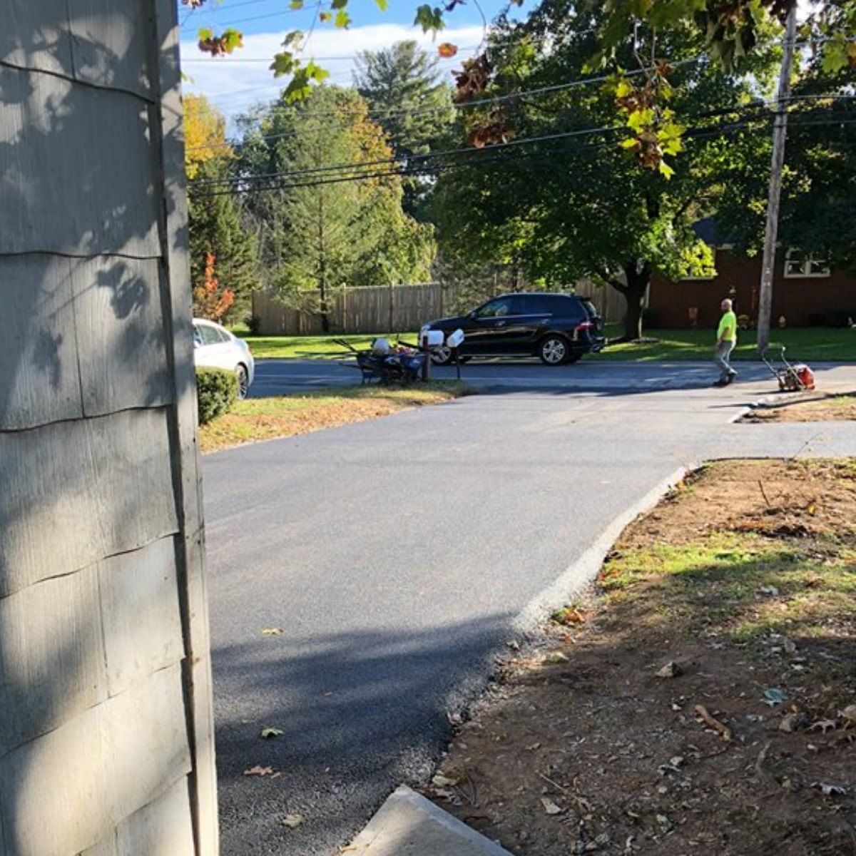 Asphalt driveway being paved; worker operates equipment. A black SUV and white car are parked nearby.