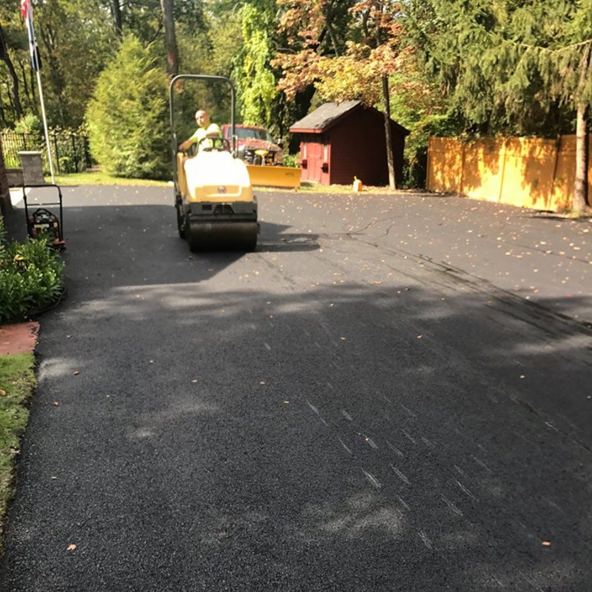 Person operating a road roller on freshly paved asphalt driveway, with a small shed and trees in the background.