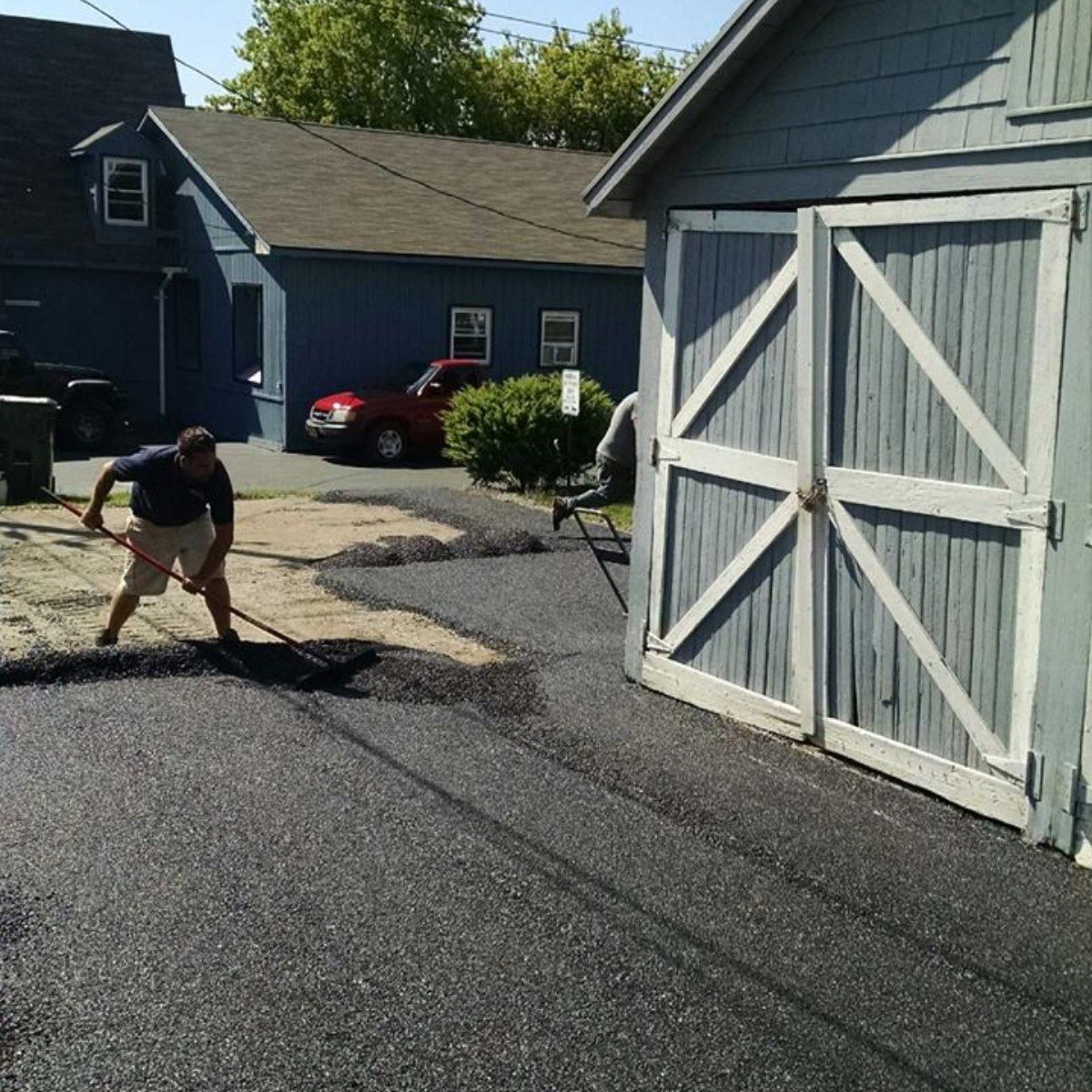 Man raking asphalt on a driveway next to a blue garage and houses.