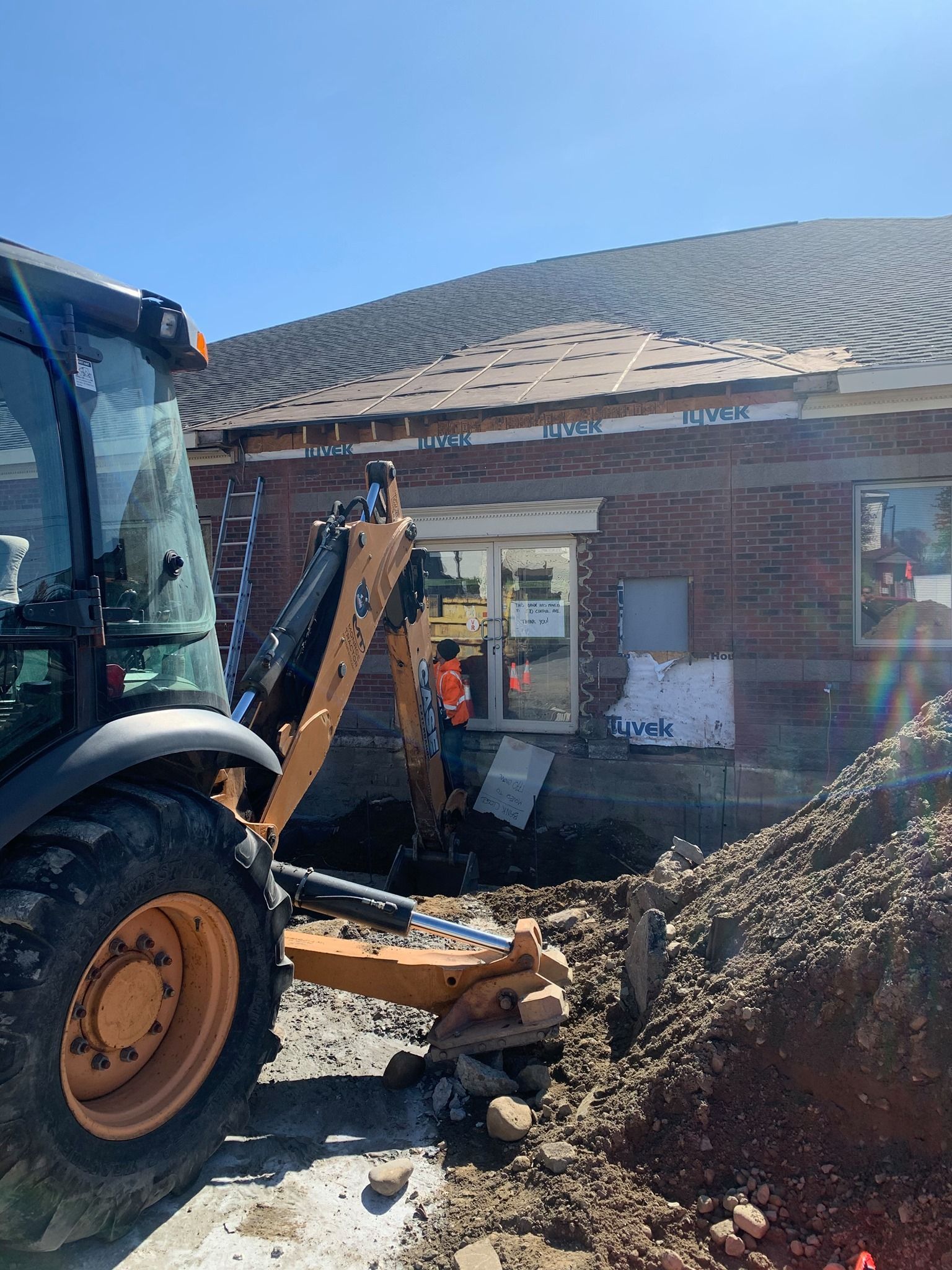 Construction site with a backhoe digging near a brick building with a partially exposed roof under a blue sky.