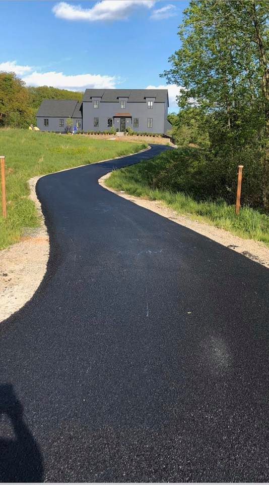 Paved black driveway curves up to a gray house on a sunny day. Green grass and trees line the sides.