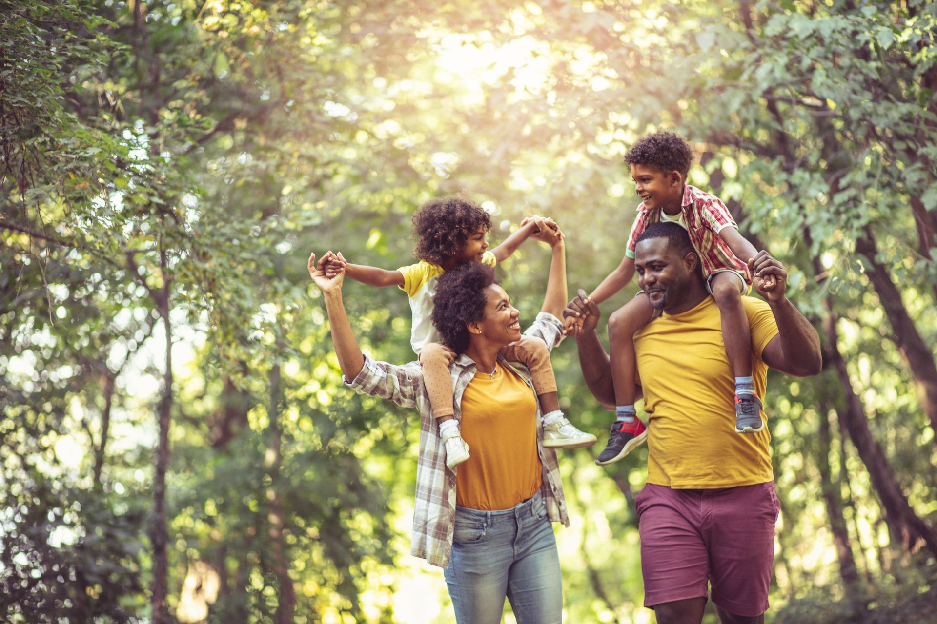 Family of four walking in a sunny park; parents carrying children on their shoulders.