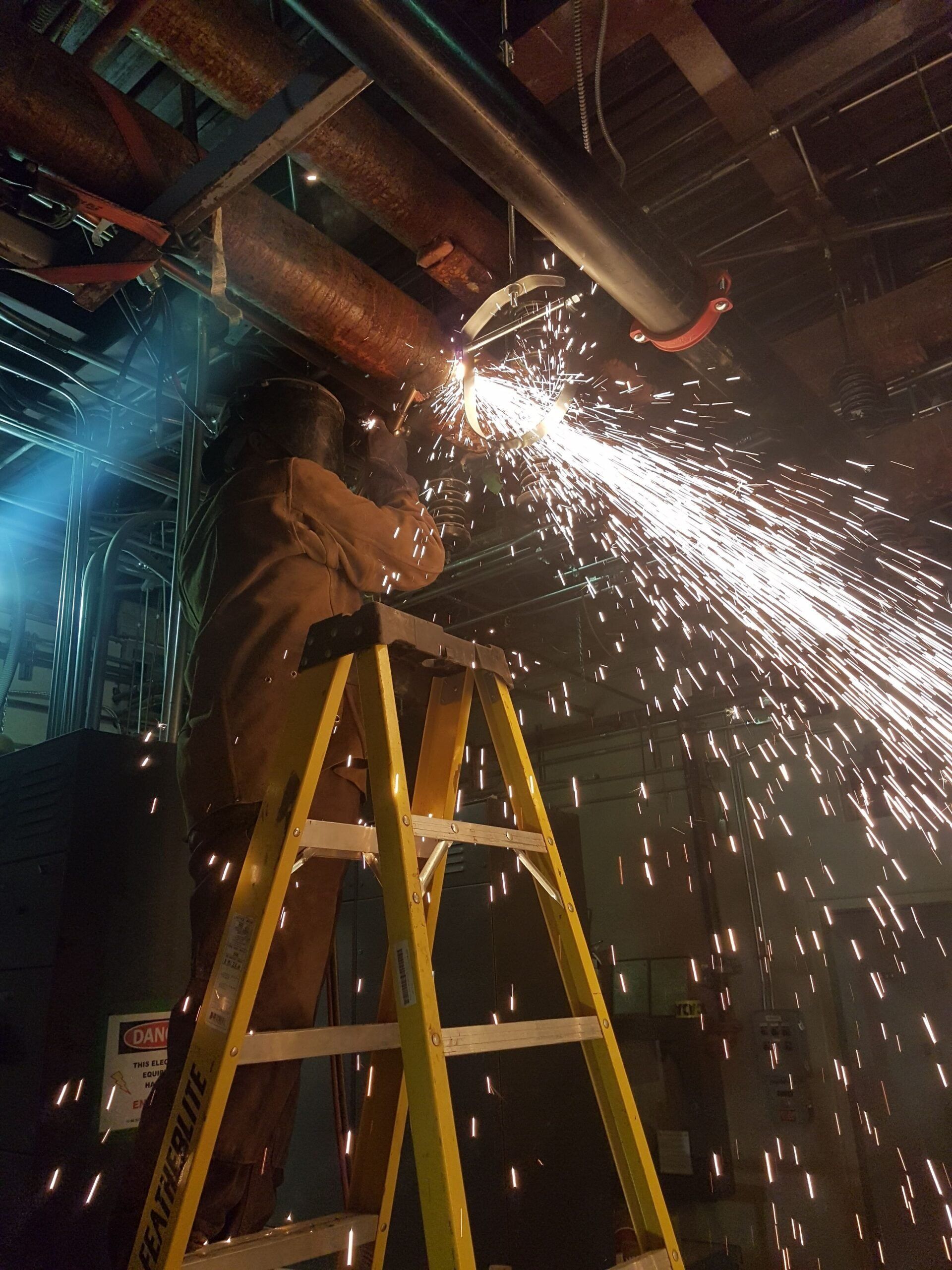 A man is standing on a ladder cutting a pipe.