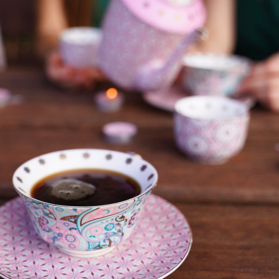 A close-up of a floral teacup with a tea bag, set on a saucer, with a teapot and hands in the blurry background — Yoni Steam Australia in Gold Coast, QLD