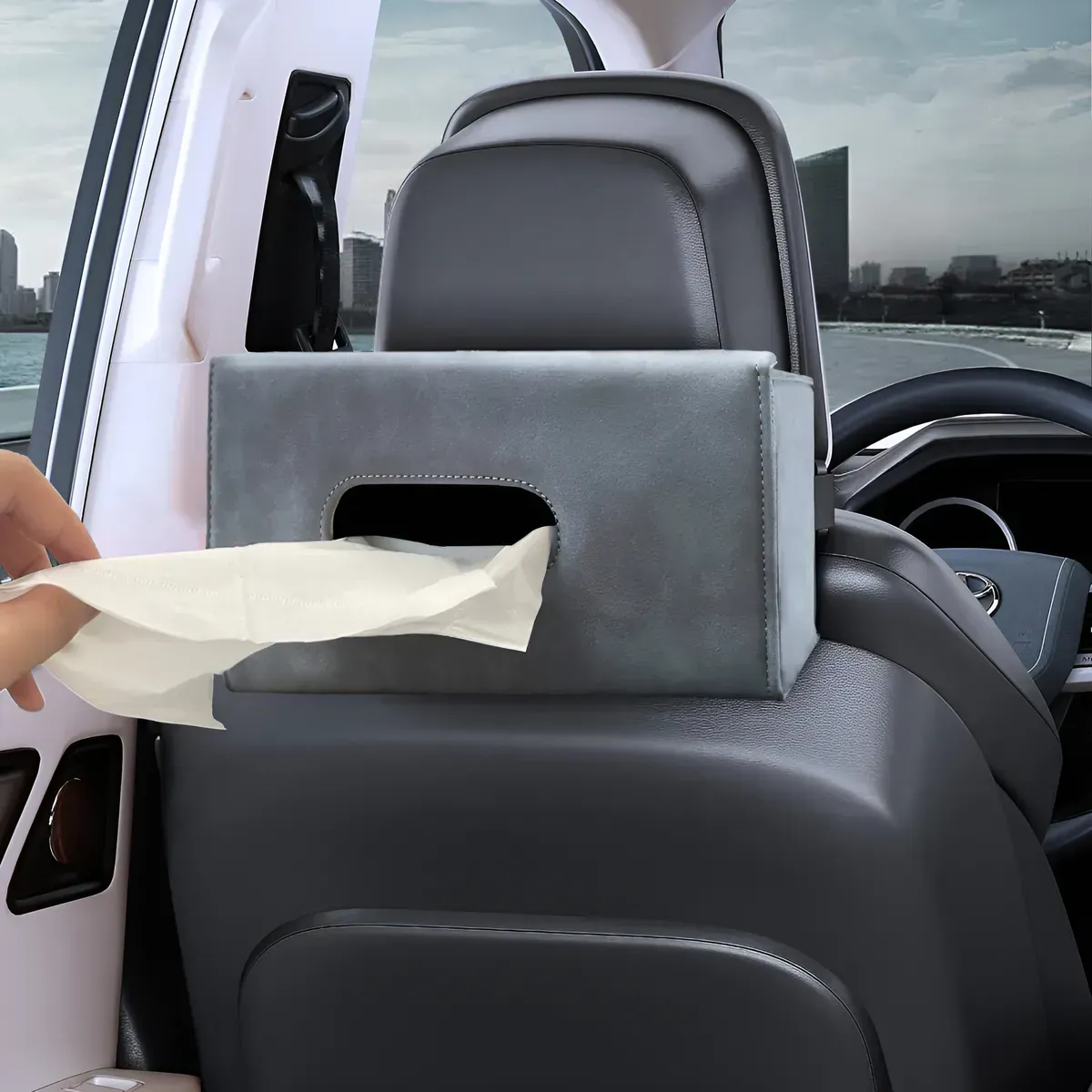 Caja de pañuelos gris en el respaldo del asiento de un automóvil, una mano sacando un pañuelo, vista de la ciudad de fondo.