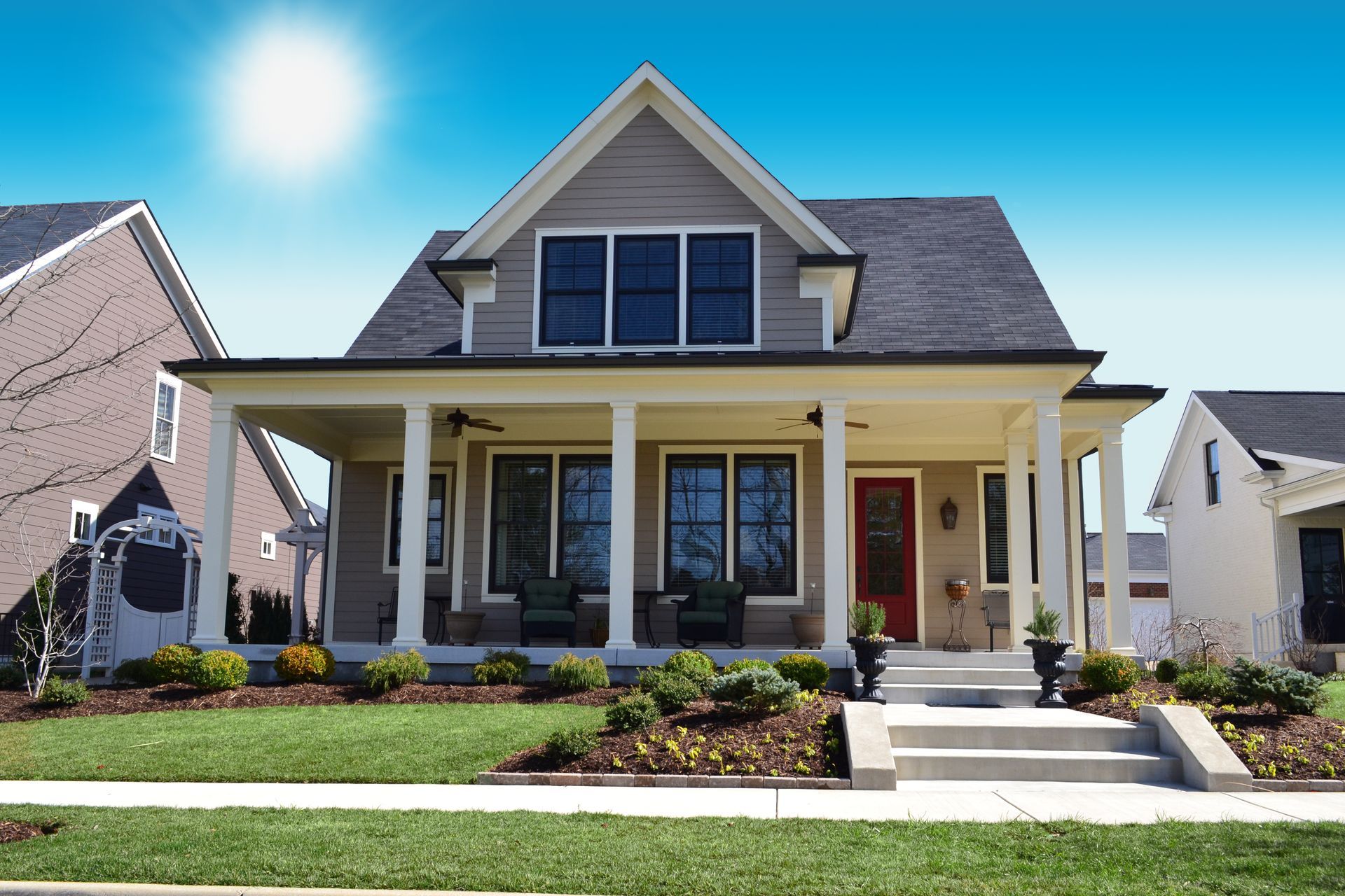 Modern suburban house with a front porch, gray siding, red door, and landscaped yard in bright sunlight