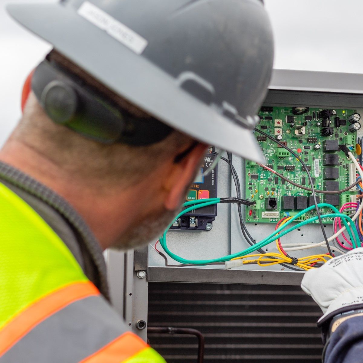 Technician in hard hat and safety vest inspecting electrical panel with circuit board and wires.