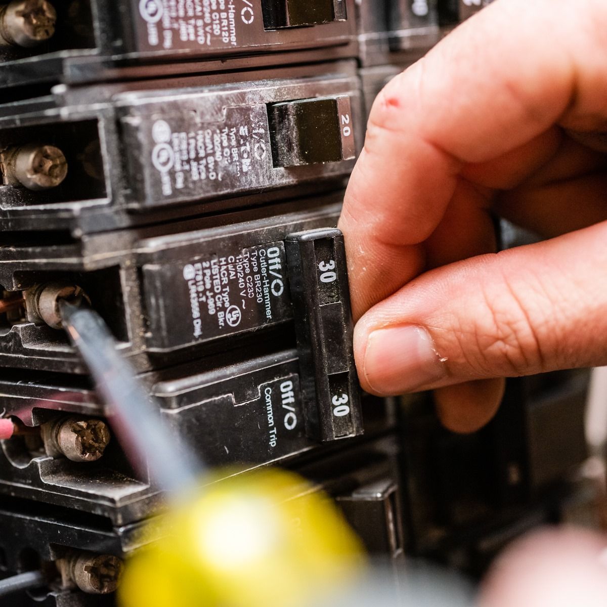 Hand turning off a 30-amp circuit breaker in a panel. Black circuit breakers with a black and gray screwdriver.
