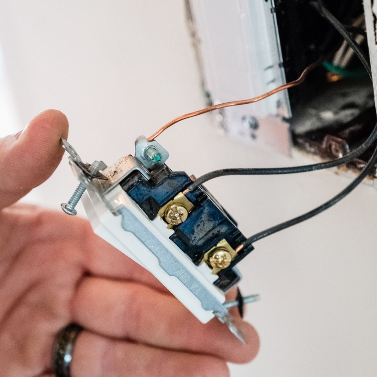 Hand holding a light switch with black and copper wires; installation on white wall.