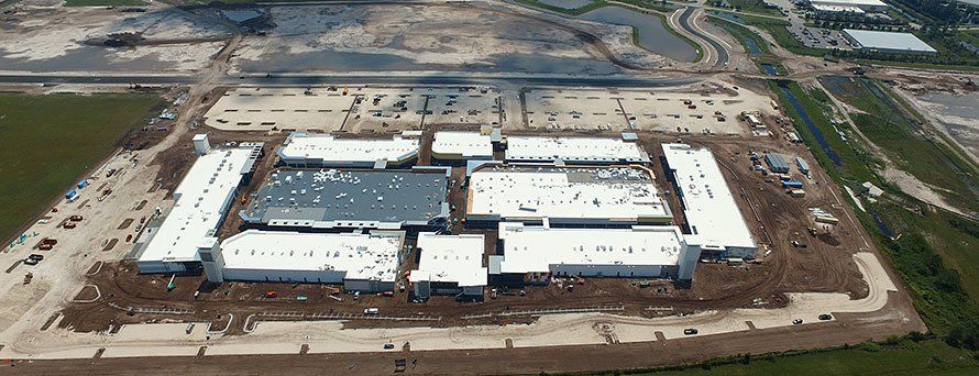 Aerial view of a large commercial building under construction with several white roofs and surrounding cleared land.