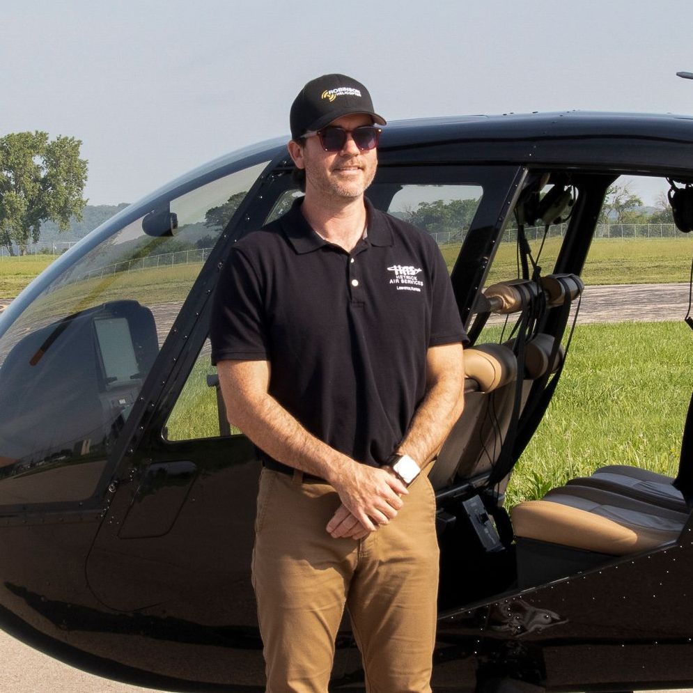 A man wearing a hat and sunglasses stands in front of a helicopter