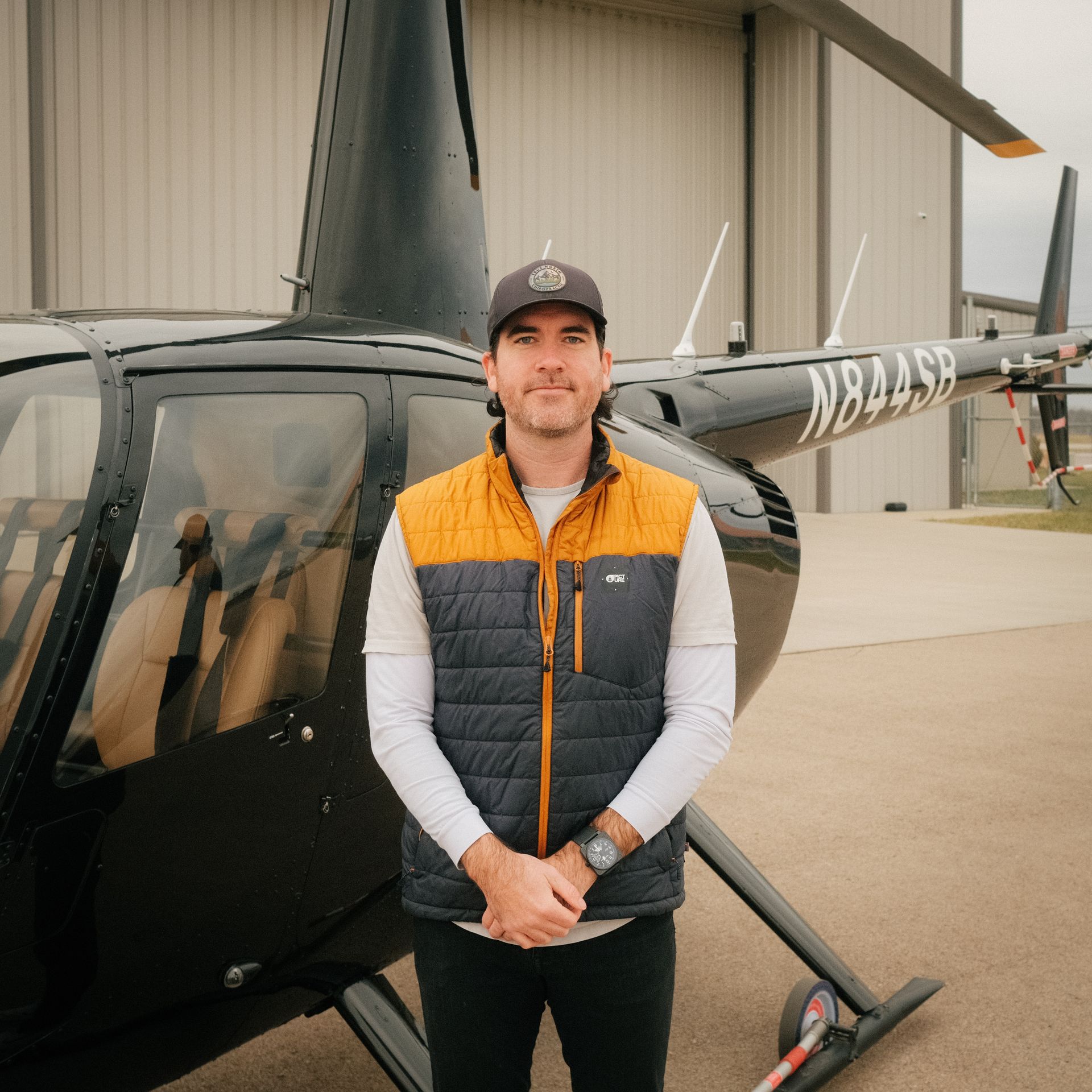 A man wearing a hat and sunglasses stands in front of a helicopter