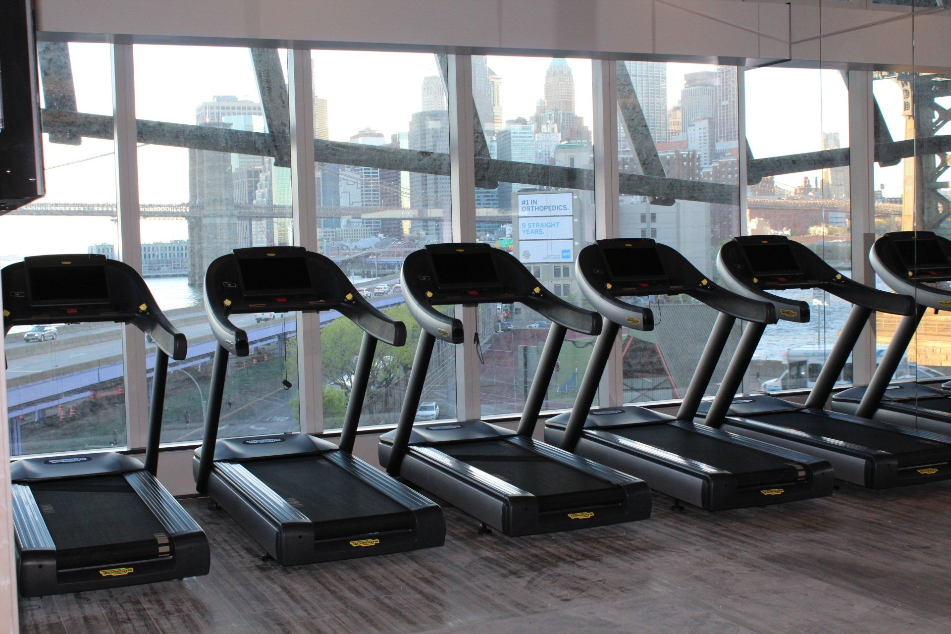 Modern treadmills lined up with panoramic views of Manhattan skyline through glass walls.