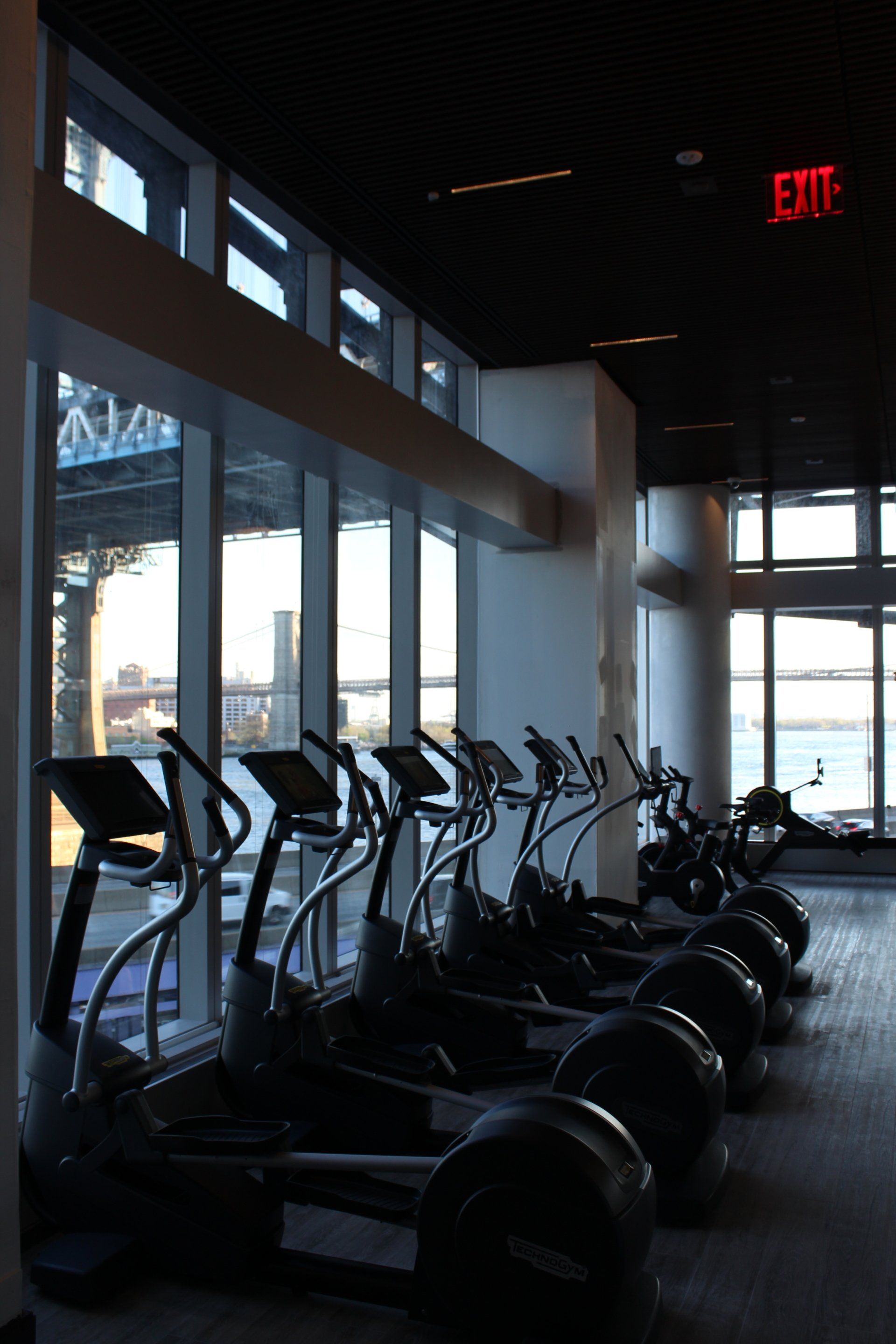 Row of cardio machines in luxury gym overlooking Manhattan Bridge and city waterfront.